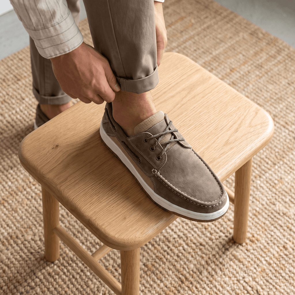 Man in cuffed pants resting his foot on a wooden stool while wearing a gray suede Adrian boat shoe, on a jute rug