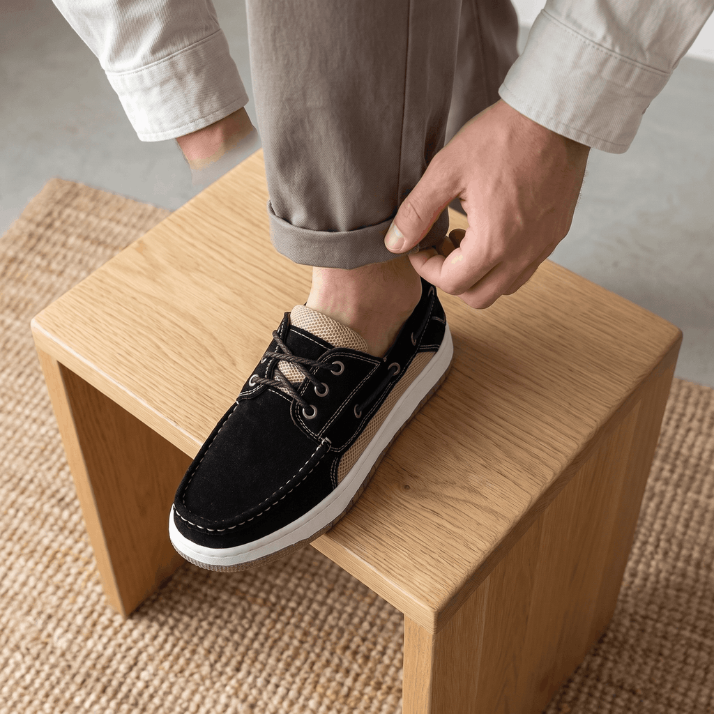 Man in cuffed trousers resting his foot on a wooden block while adjusting his black suede Adrian boat shoe, on a jute rug