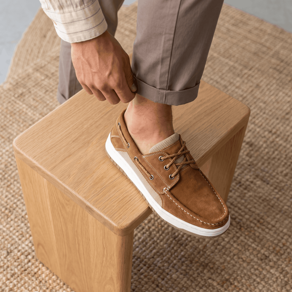 Man in rolled-up trousers resting his foot on a wooden block while adjusting his tan suede Adrian boat shoe, standing on a jute rug