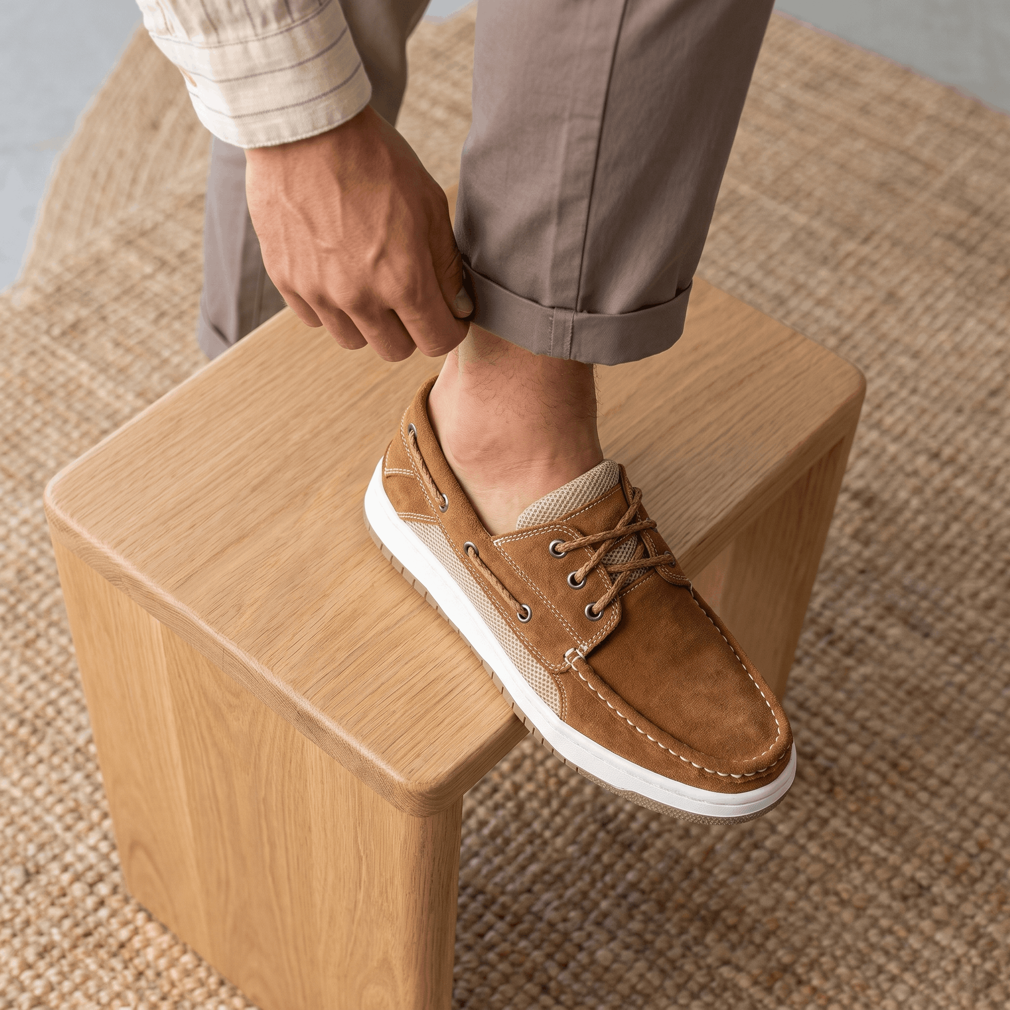Man in rolled-up trousers resting his foot on a wooden block while adjusting his tan suede Adrian boat shoe, standing on a jute rug