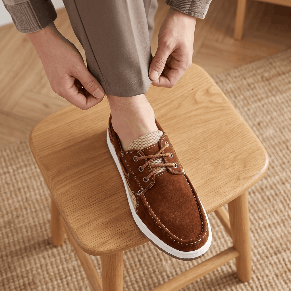 Man in trousers resting his foot on a wooden stool while adjusting his dark brown suede Adrian boat shoe, standing on a jute rug