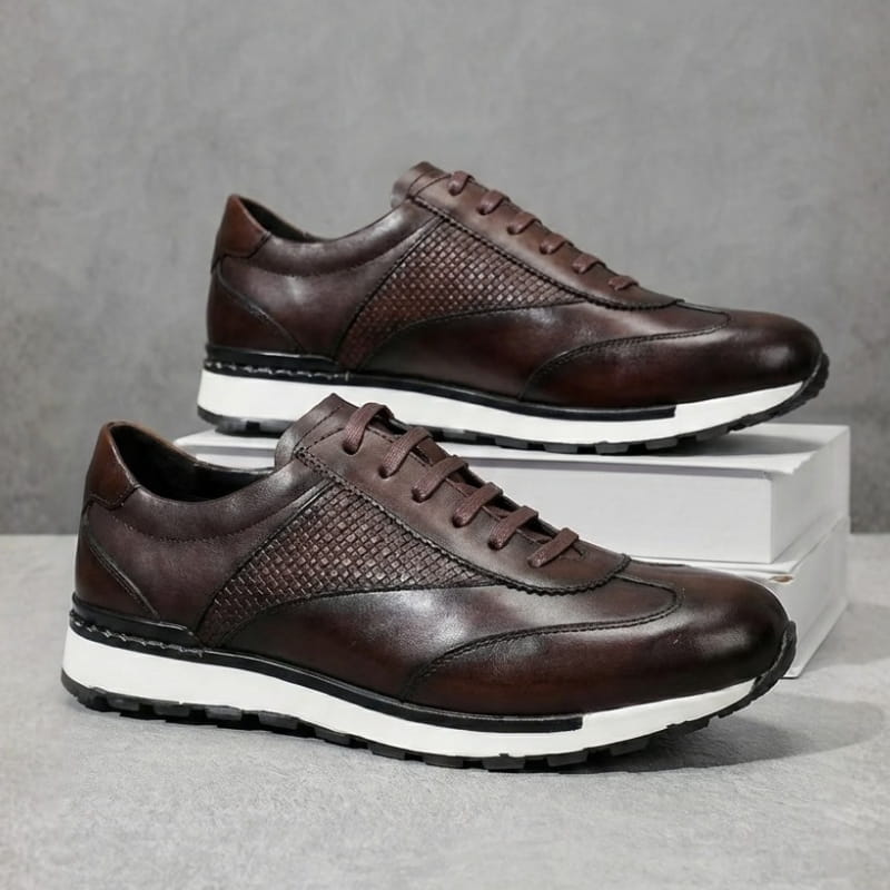 Pair of dark brown leather sneakers with woven texture panels, stacked on a light shoebox against a gray background.