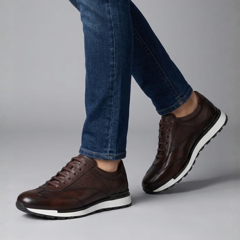 A person walking wearing dark brown leather sneakers and blue jeans against a plain gray background.