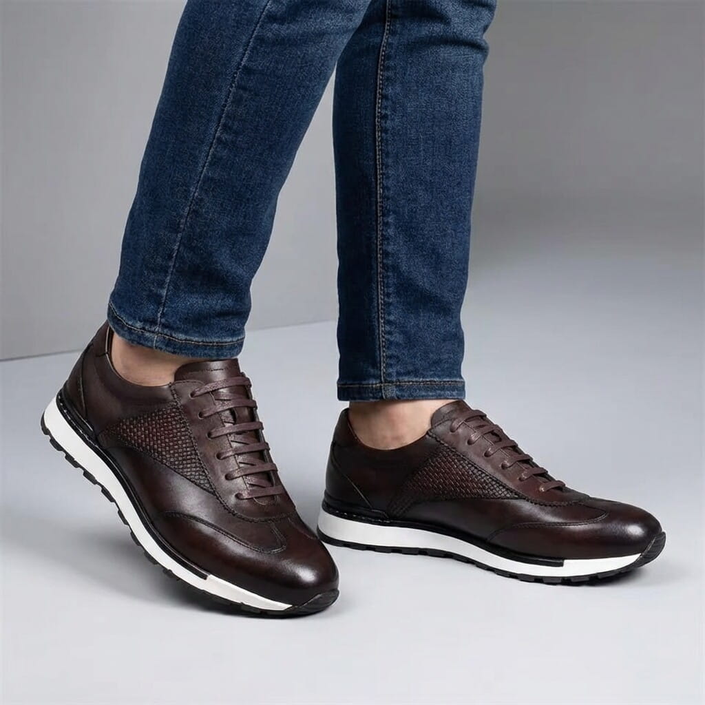 A person standing in dark brown leather sneakers and blue jeans against a gray background.