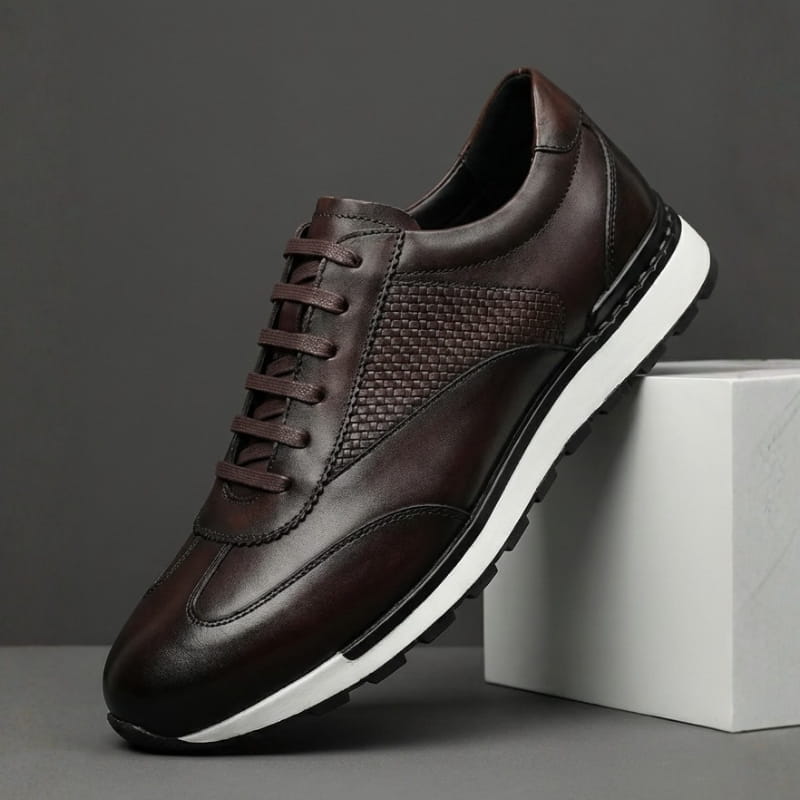 A single dark brown leather sneaker with a woven texture panel and white sole, propped against a concrete block.