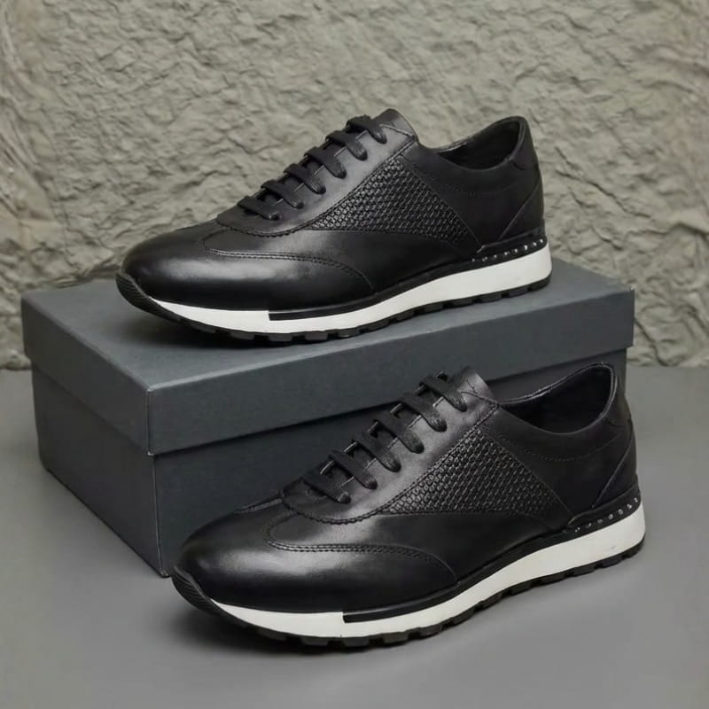 Pair of black leather sneakers with woven texture panels, stacked on a gray shoebox against a textured wall.