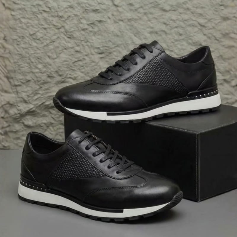 Pair of black leather sneakers with woven texture panels, resting beside a dark shoebox against a textured wall.