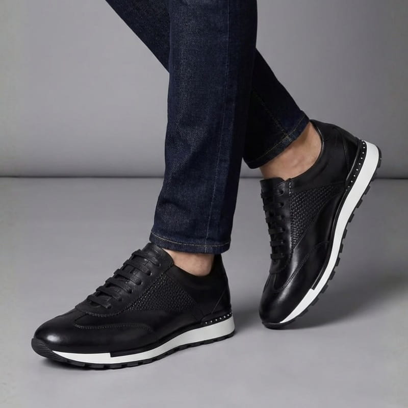 A person's legs crossed mid-step wearing black leather sneakers with perforated detailing and cuffed dark denim jeans.