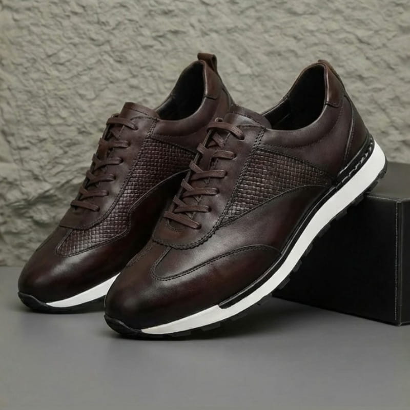 Pair of dark brown leather sneakers with woven texture panels, resting against a dark box against a textured wall.