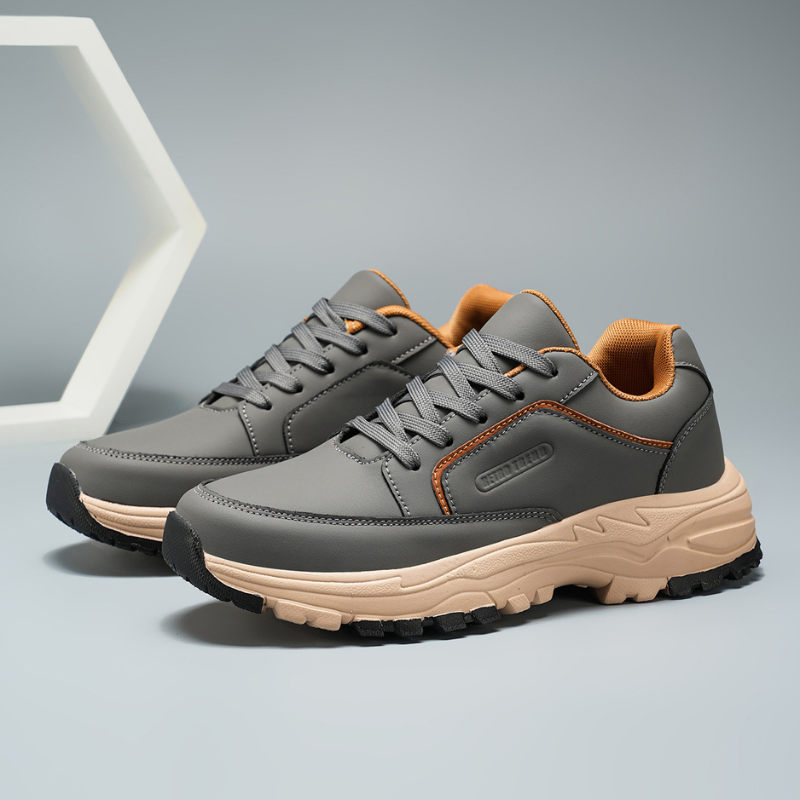 Front-facing view of gray leather Arnold trainers with orange lace and collar accents, set against a geometric hexagon backdrop