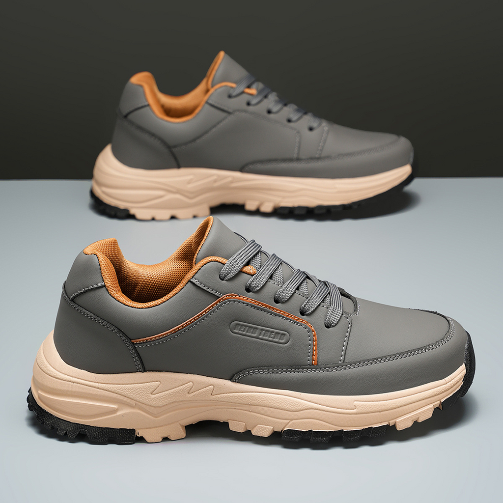 Gray leather Arnold trainers with orange trim tilted on their side against a two-tone olive and gray backdrop, showing the tan tread sole