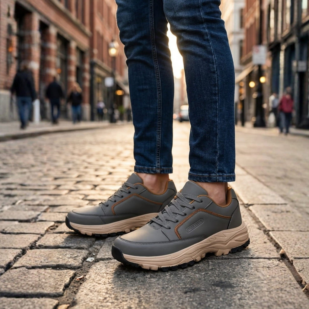 Man wearing gray leather Arnold trainers with orange trim and cuffed jeans, standing on a cobblestone city street lined with brick buildings
