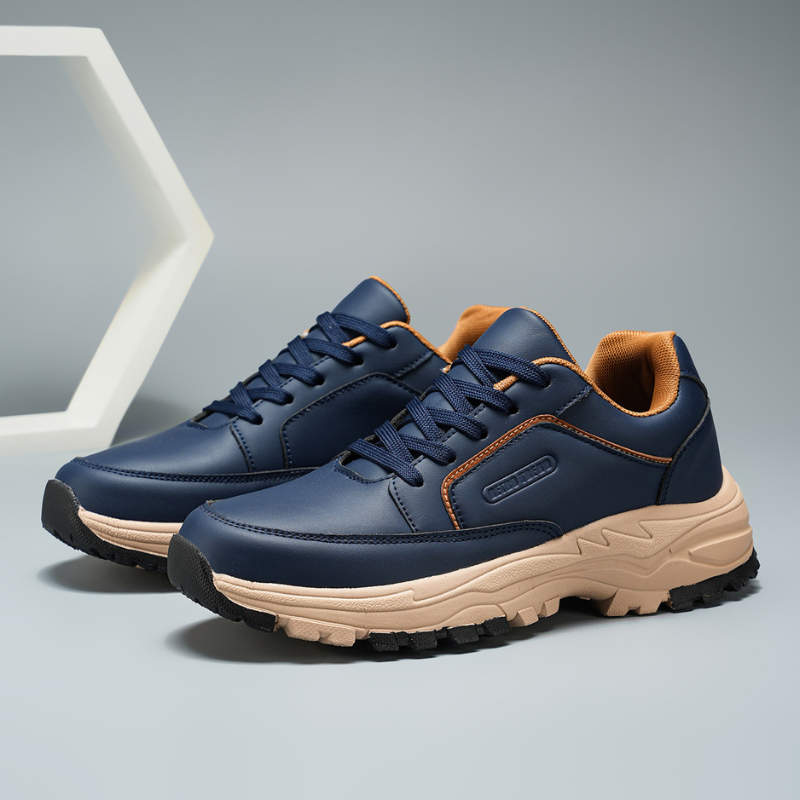 Front-facing view of navy blue Arnold trainers with orange lace and collar accents, set against a geometric hexagon backdrop