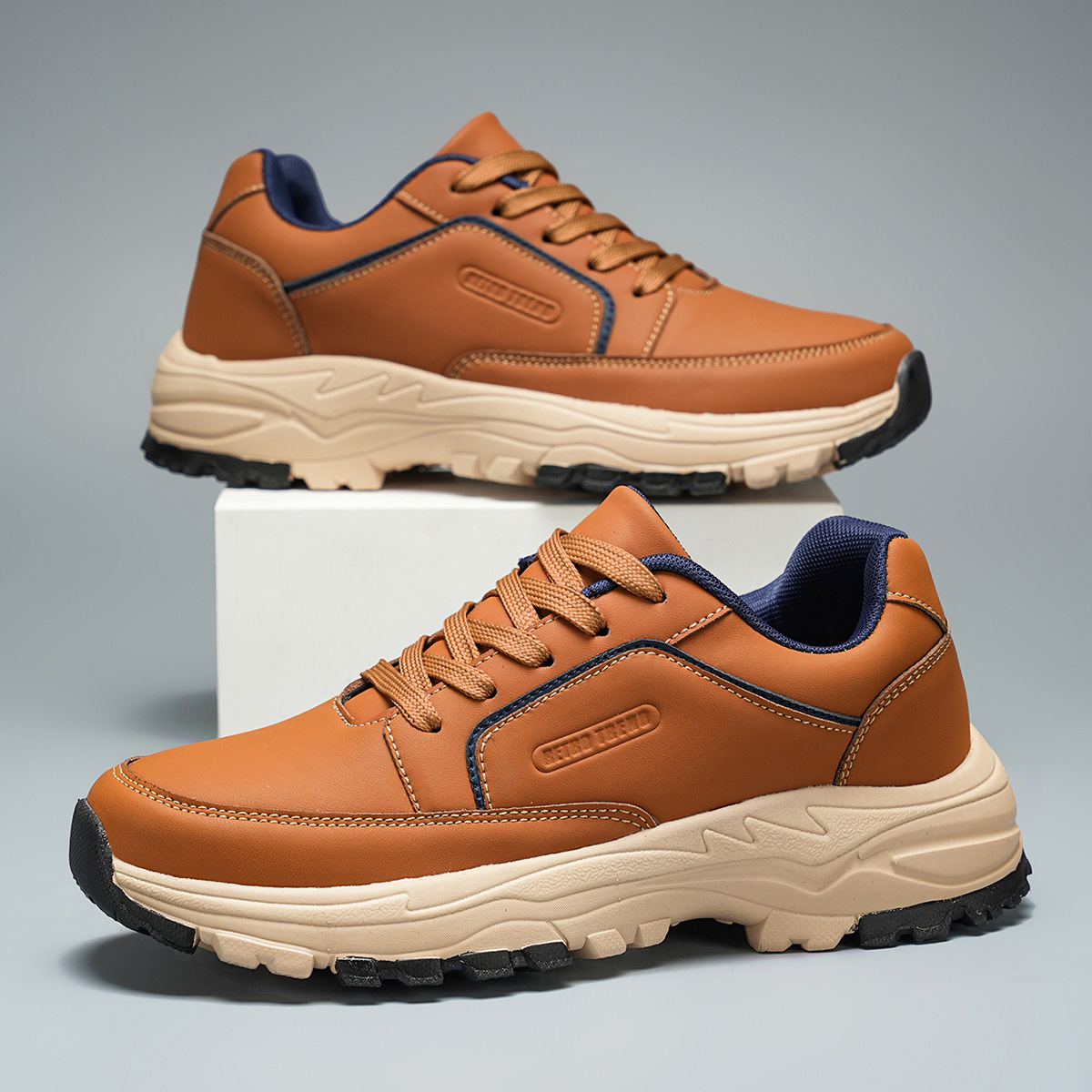 Pair of tan leather Arnold trainers with navy trim and cream sole, one shoe propped upright on a white block against a gray background