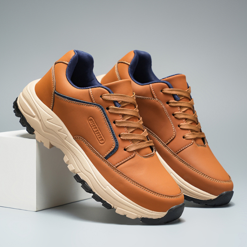 Close-up of two tan leather Arnold trainers with navy collar lining leaning together against a white block on a gray backdrop