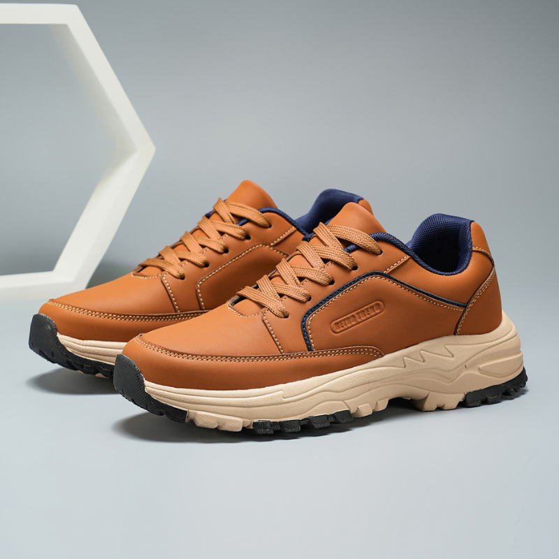 Tan leather Arnold trainers with navy trim shown at an angle beside a white geometric hexagon prop on a gray backdrop
