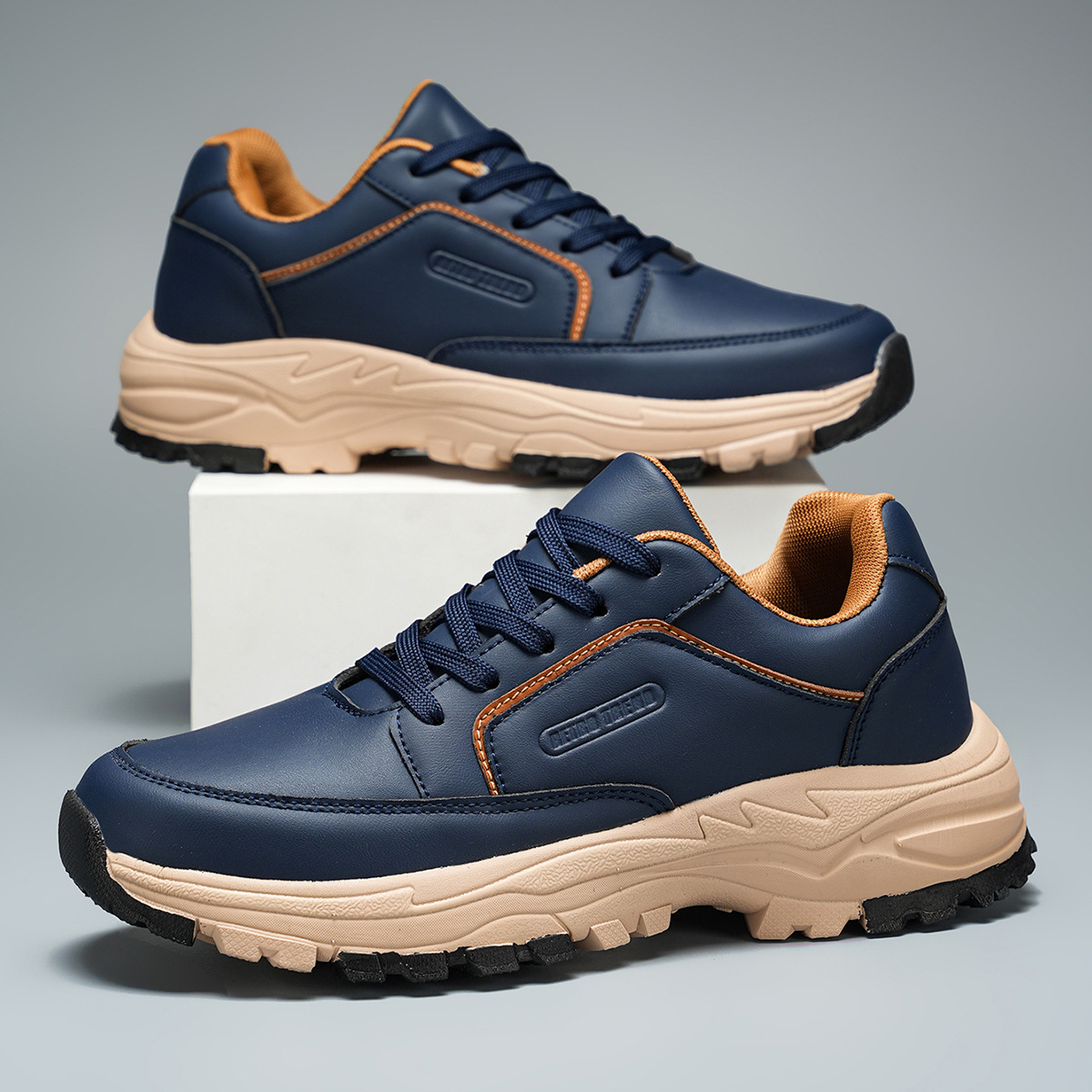 Pair of navy blue leather Arnold trainers with tan cushioned sole and orange trim accents, one shoe propped upright on a white block