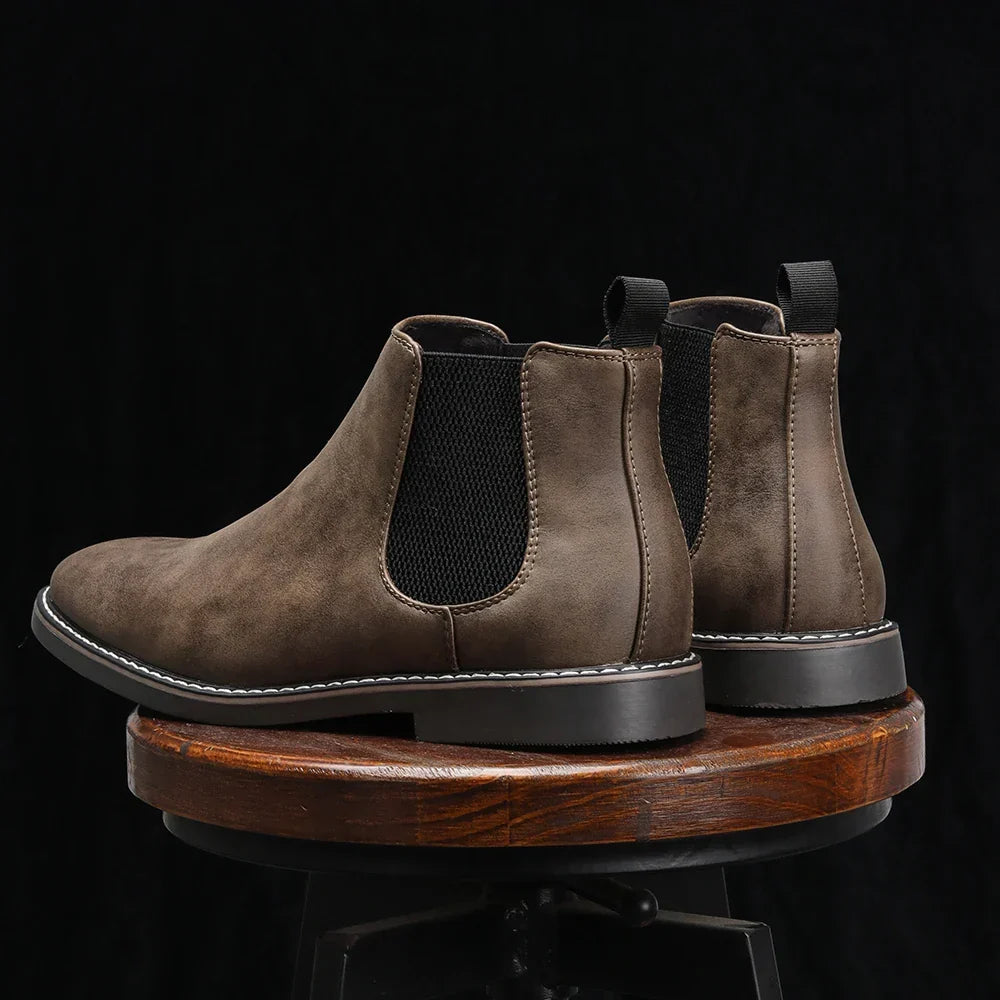 Rear view of taupe full-grain leather Chelsea boots with pull tabs and elastic side panels, resting on a wooden stool against a dark background.