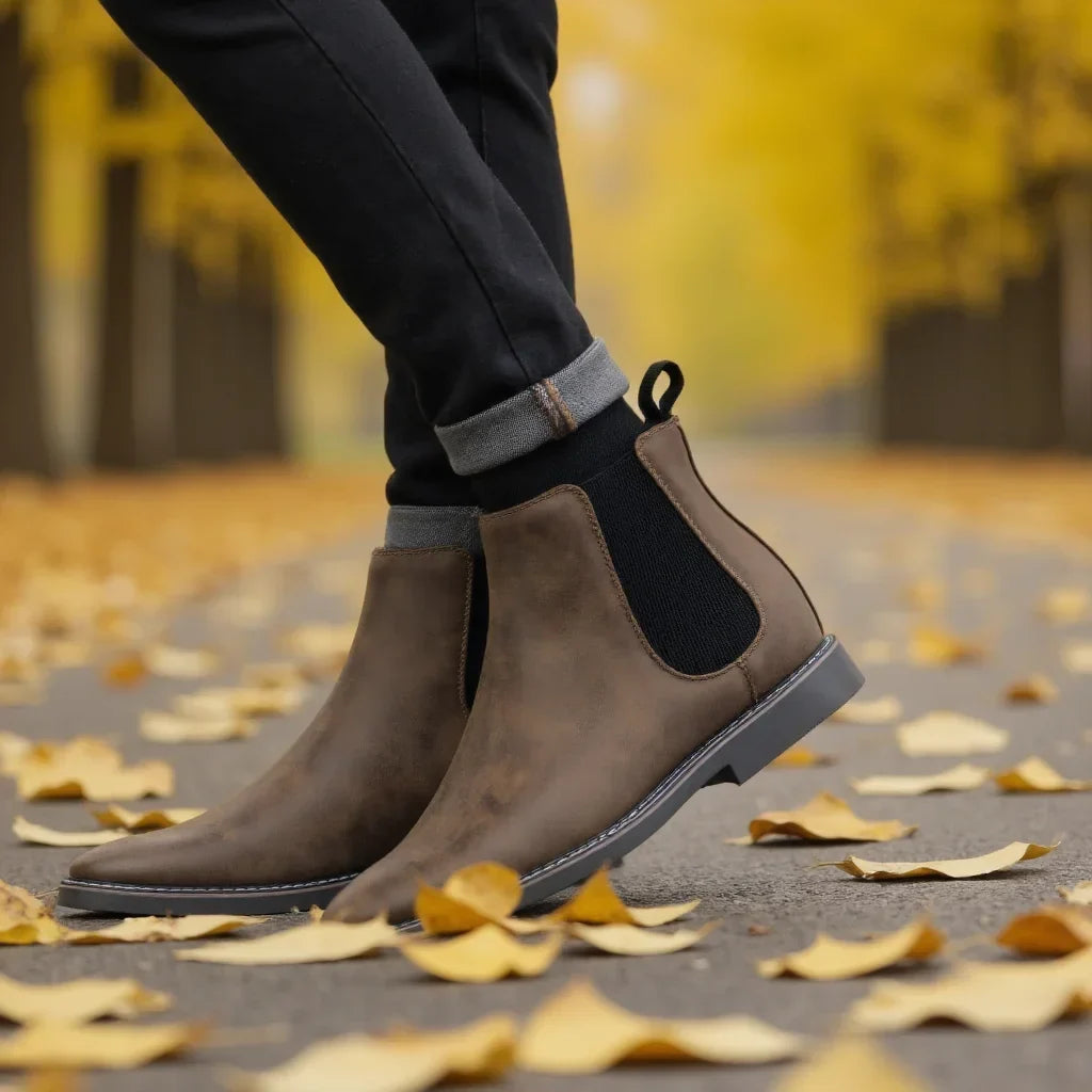 A model's feet in cuffed dark trousers wearing taupe leather Chelsea boots, walking along a path covered in fallen autumn leaves.