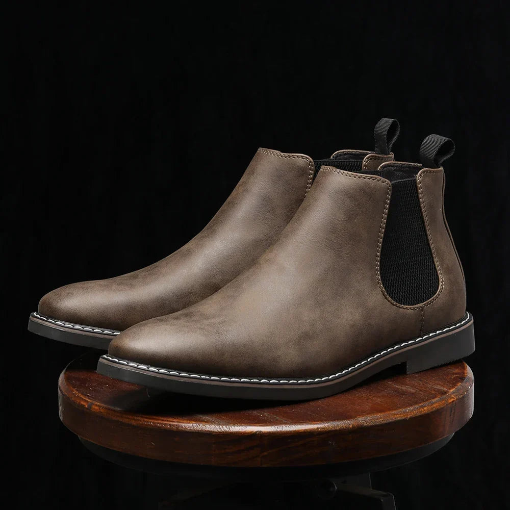 Pair of taupe full-grain leather Chelsea boots with elastic side panels, resting on a round wooden pedestal against a dark background.