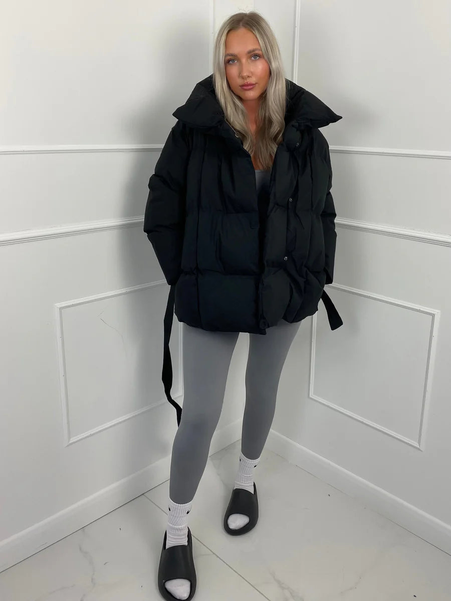 Black oversized quilted puffer jacket with a high stand collar and self-tie waist belt worn over grey leggings.
