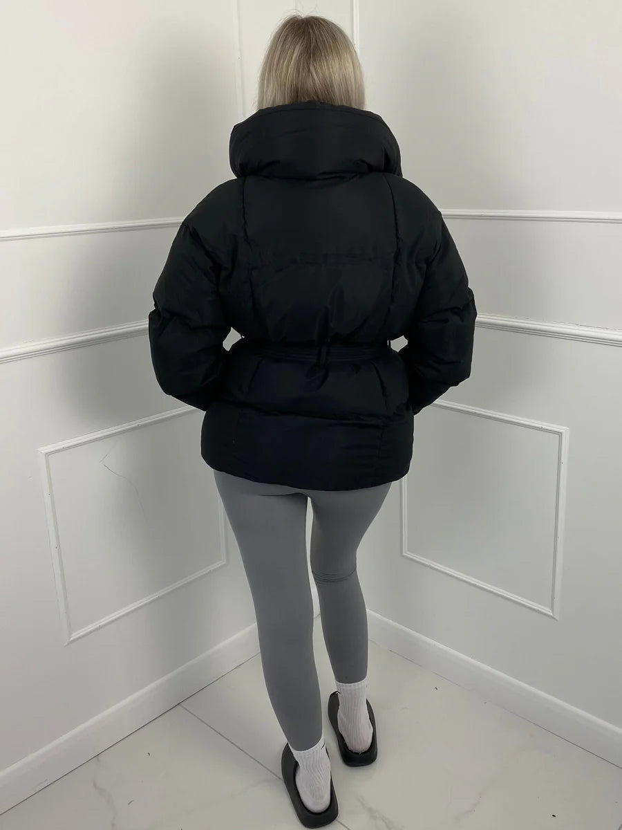 Black quilted puffer jacket with an exaggerated stand collar and self-tie waist belt, worn over grey leggings.