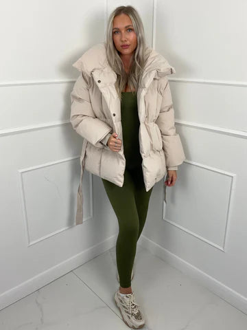 Oversized beige quilted puffer jacket with a high stand collar and self-tie waist belt worn over an olive jumpsuit.