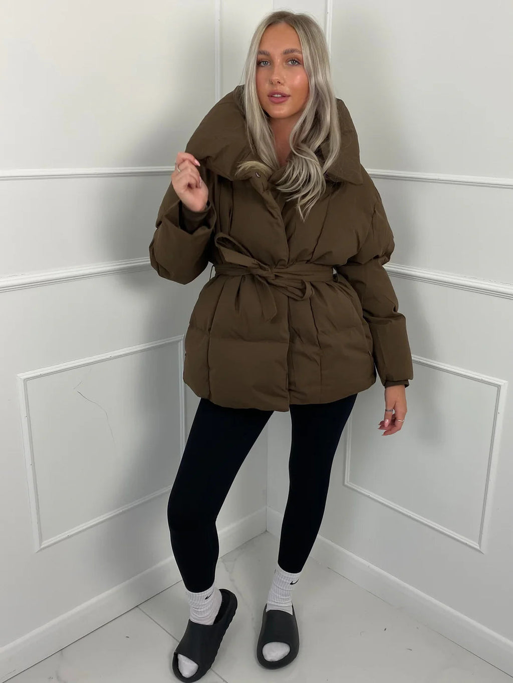 Brown oversized puffer jacket with a high stand collar and self-tie waist belt worn over black leggings.