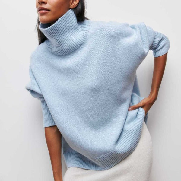 Woman wearing a light blue oversized ribbed turtleneck sweater with short sleeves and a relaxed fit paired with white bottoms.