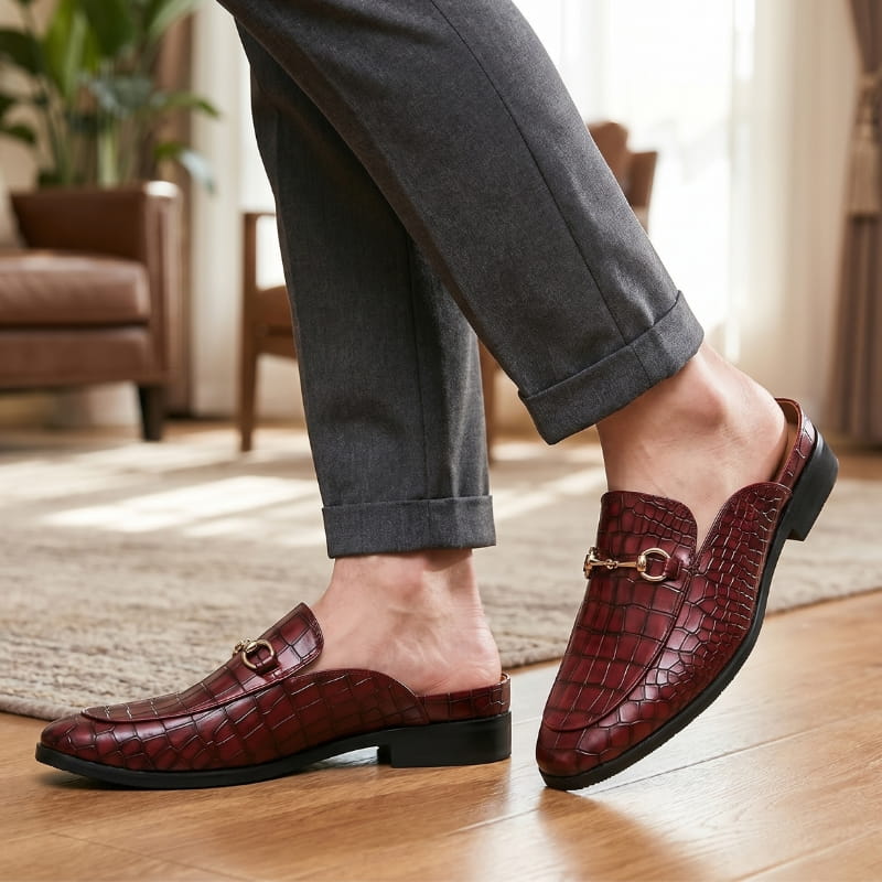 Legs in cuffed gray trousers wearing burgundy croc-embossed mule loafers, walking on a wooden floor with a jute rug
