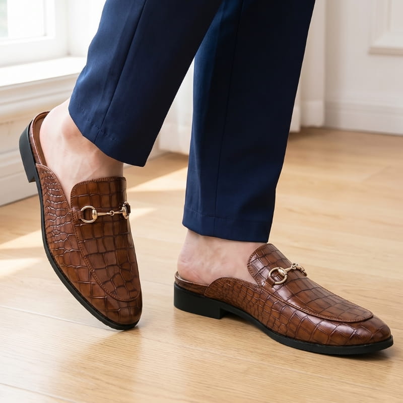 Legs in navy trousers wearing brown croc-embossed mule loafers, standing on a wooden floor near a window