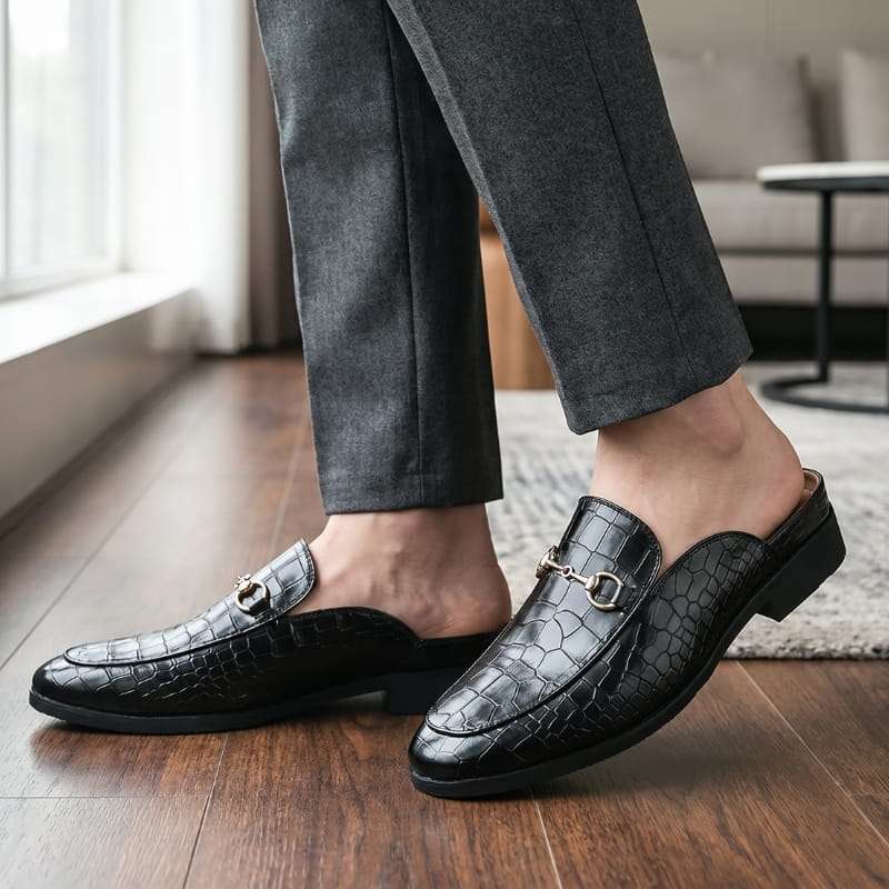 Legs in gray trousers wearing black croc-embossed mule loafers, standing on a wooden floor beside a sunlit window