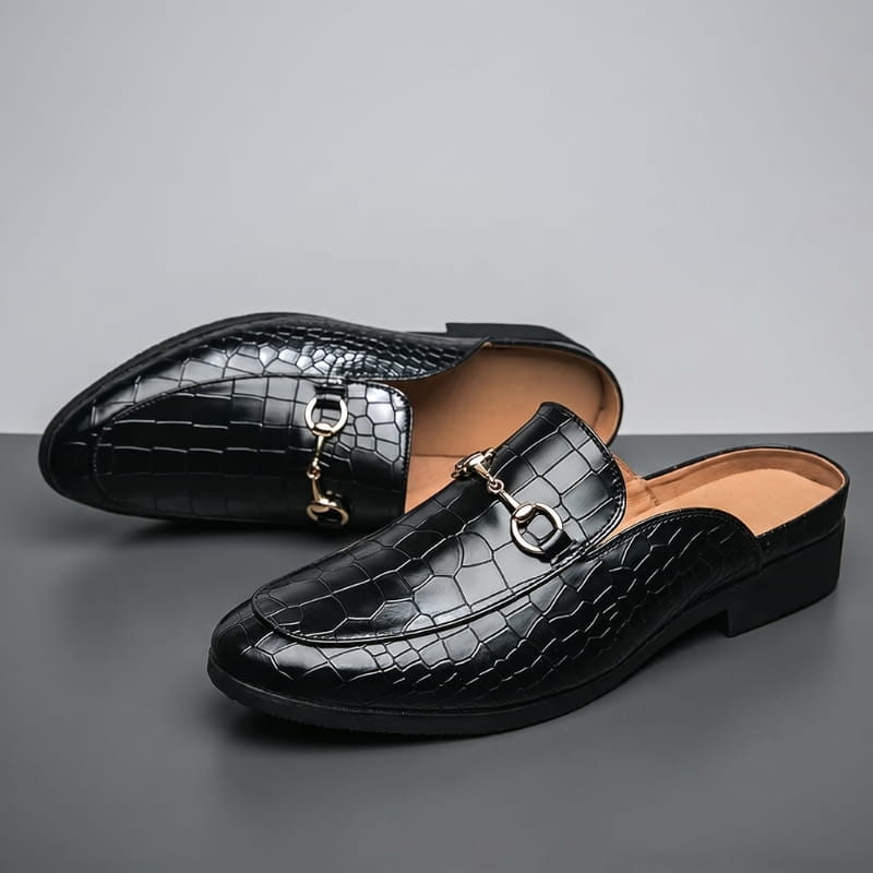 Pair of black croc-embossed leather mule loafers with gold horsebit buckles, one shoe tipped on its side, on a gray backdrop