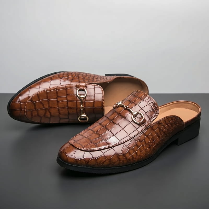 Pair of brown croc-embossed leather mule loafers with gold horsebit buckles, one shoe tipped on its side, on a gray backdrop