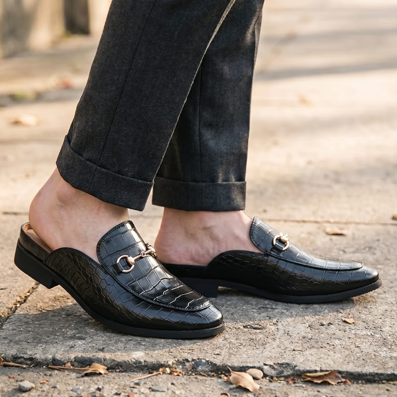Bare ankles in cuffed dark trousers wearing black croc-embossed mule loafers, standing on outdoor stone pavement