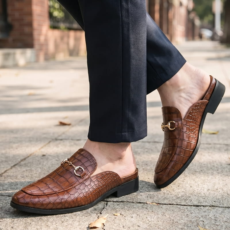 Bare ankles in cuffed navy trousers wearing brown croc-embossed mule loafers, walking on an outdoor stone path
