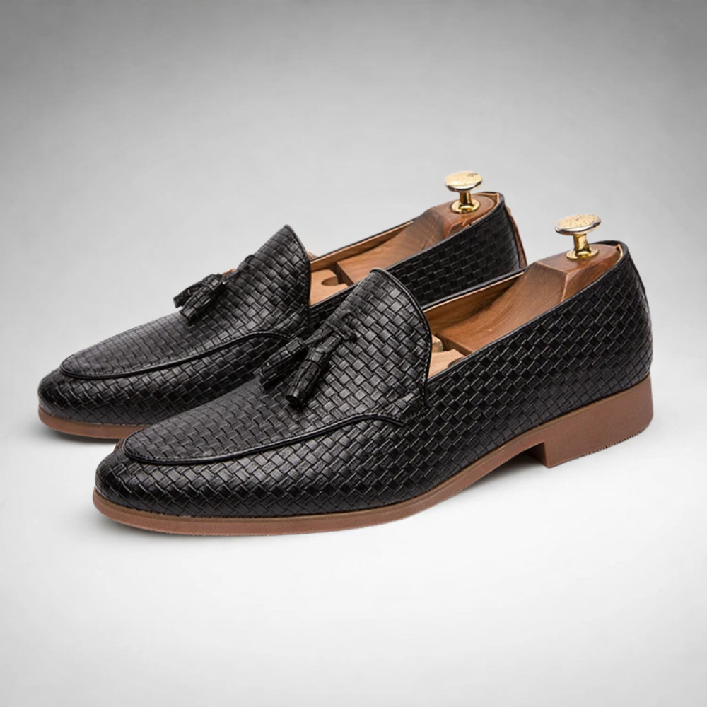 Pair of black woven leather tassel loafers with wooden shoe trees inside, studio product shot on a light gray background.