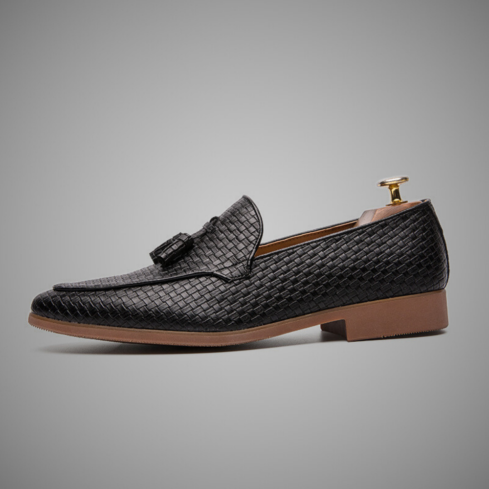 Side profile product shot of a black woven leather tassel loafer with a wooden shoe tree, on a light gray background.