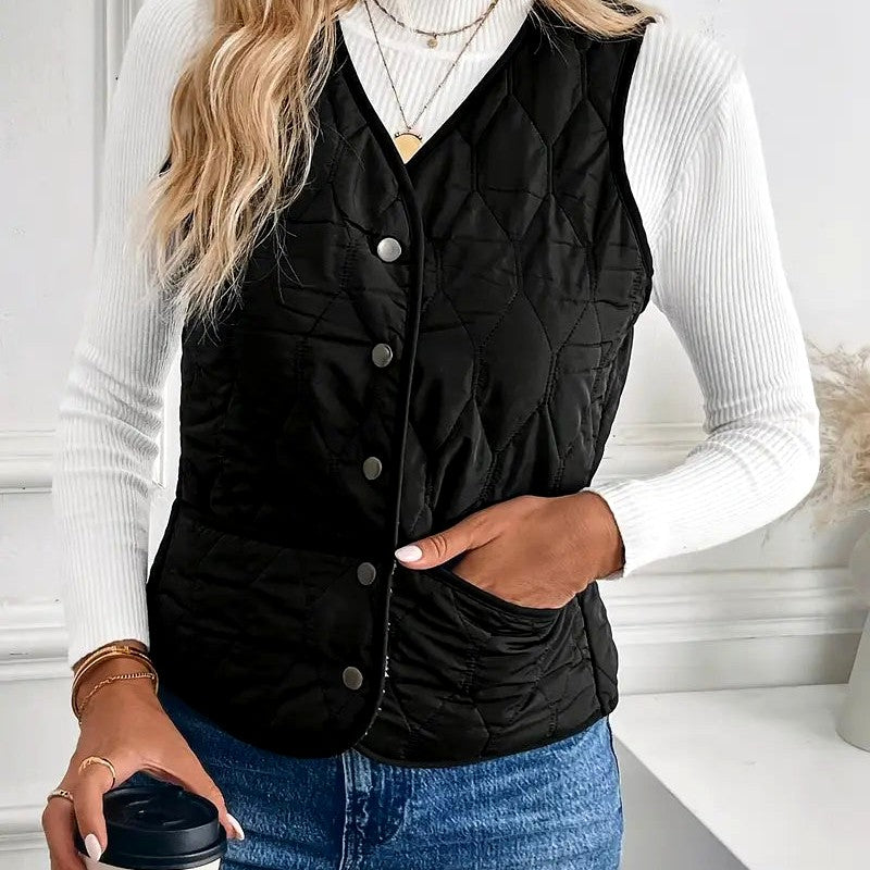 Woman wearing a black quilted puffer vest with a stand collar, snap-button front, and diamond quilted finish over a white ribbed sweater.