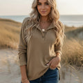Woman wearing a khaki waffle knit Henley sweater with a collared button neckline, long sleeves, and a relaxed fit in a beachside setting.