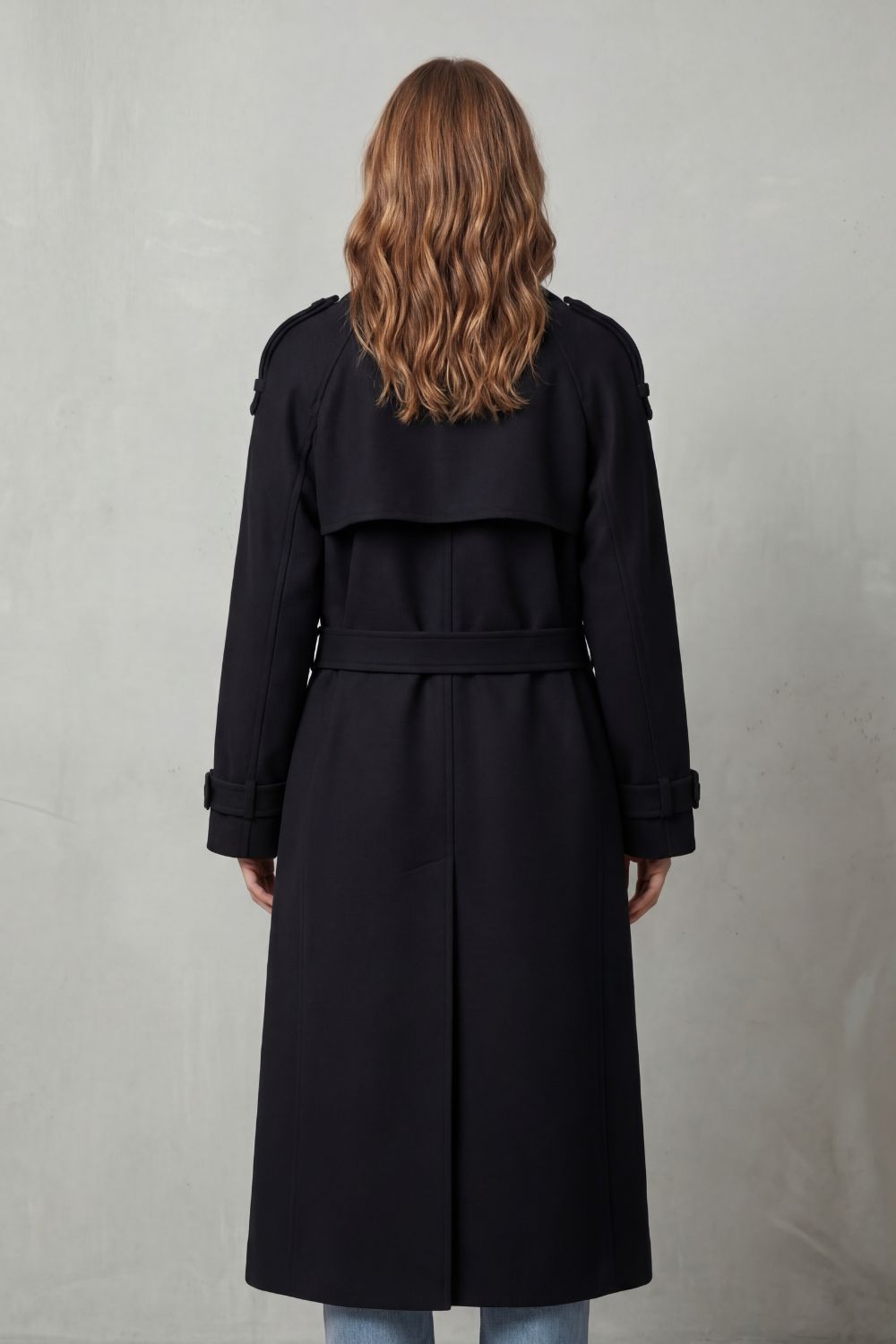 Black wool trench coat in an oversized, longline silhouette with a belted waist and structured back storm flap.
