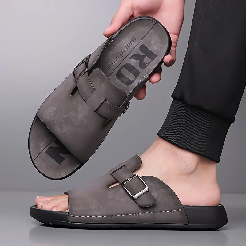 Close-up of a model's foot wearing a gray vegan leather slide sandal, with a hand adjusting the buckle strap for fit.