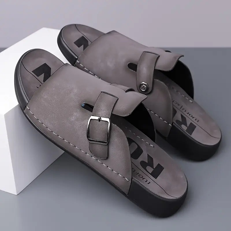 Pair of gray vegan leather slide sandals with buckle straps, styled leaning against a white geometric display prop.