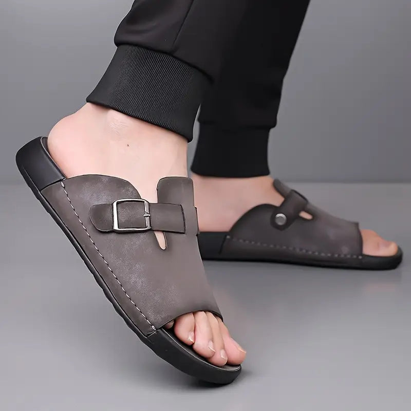 Close-up of a model's feet crossed while wearing gray vegan leather slide sandals with buckle and adjustable loop straps.