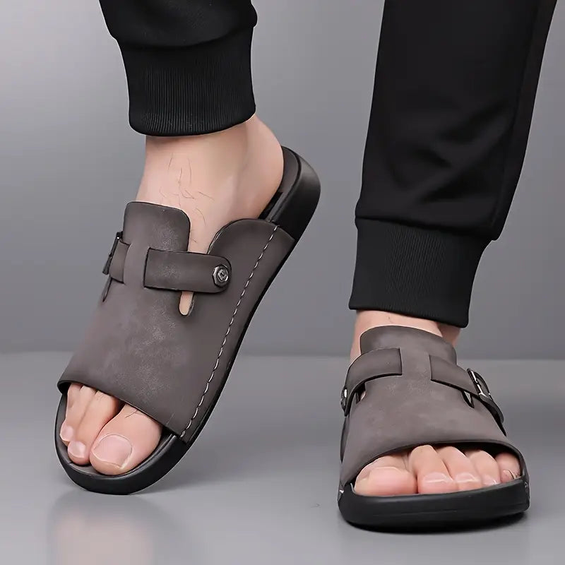 Close-up of a model's feet wearing a pair of gray vegan leather slide sandals with visible buckle straps at the ankle.