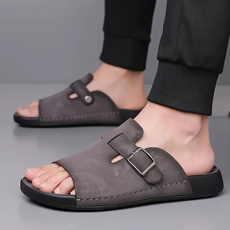Close-up angled shot of a model's foot wearing a gray vegan leather slide sandal, highlighting the buckle strap detail.