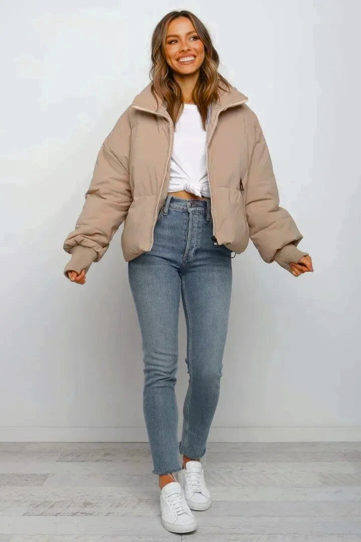 Beige cropped quilted puffer jacket with a zip front, relaxed fit, and ribbed cuffs worn over blue jeans and white sneakers.