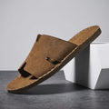 Brown suede slide sandal with H-shaped cutout strap and hand-stitched trim propped diagonally against a white geometric block