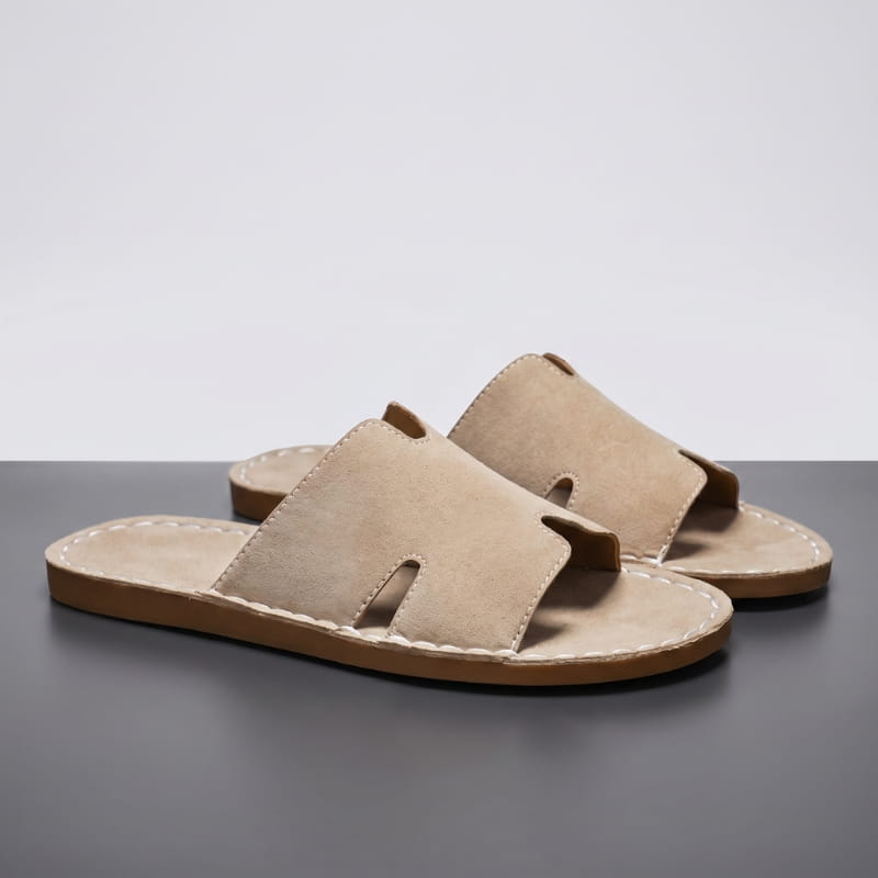Pair of khaki suede slide sandals with H-shaped cutout straps and hand-stitched trim on a gray studio floor