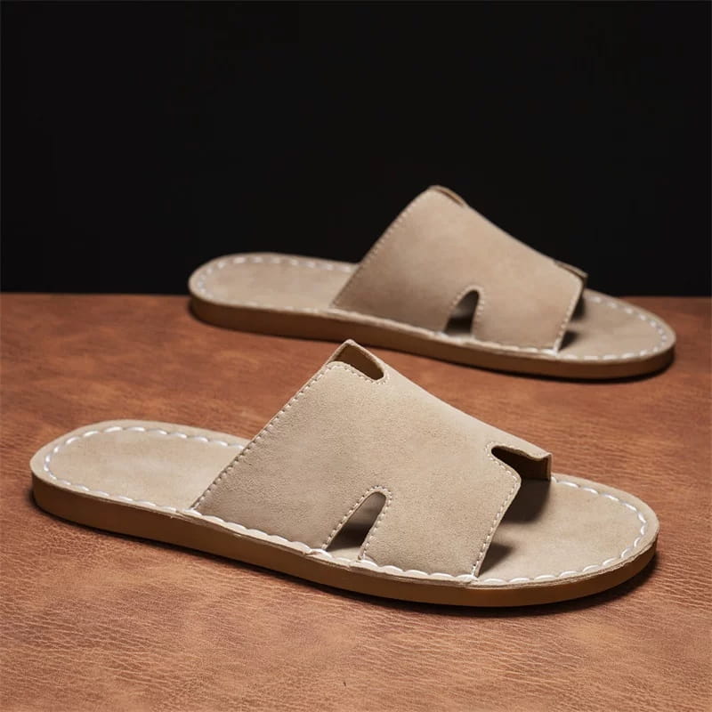 Pair of khaki suede slide sandals with H-shaped cutout straps on a warm brown leather surface with a dark backdrop